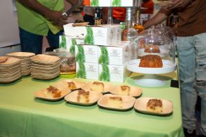 Food vendor display with baked goods on palm leaf plates, white product boxes with green palm leaf designs, glass cake dom…