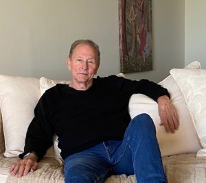Bob Lind in black sweatshirt and blue jeans sitting on white couch