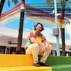 Timothy LaRoque in tie-dye shirt posing on colorful steps beneath rainbow-wrapped palm trees in outdoor plaza with gallery storefront