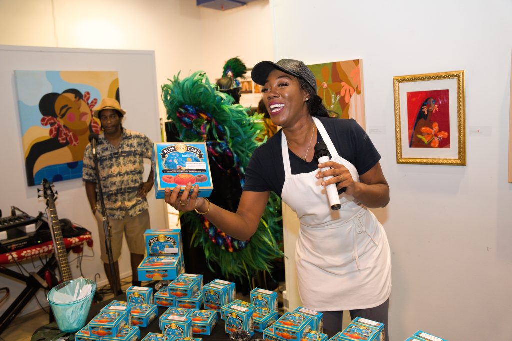Woman in white apron holding microphone and blue Rum Cake box at cultural event, with colorful artwork and performers in b…