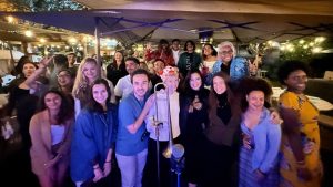 Large group of people gathered under a tent at night with string lights, posing together with a microphone for a community…